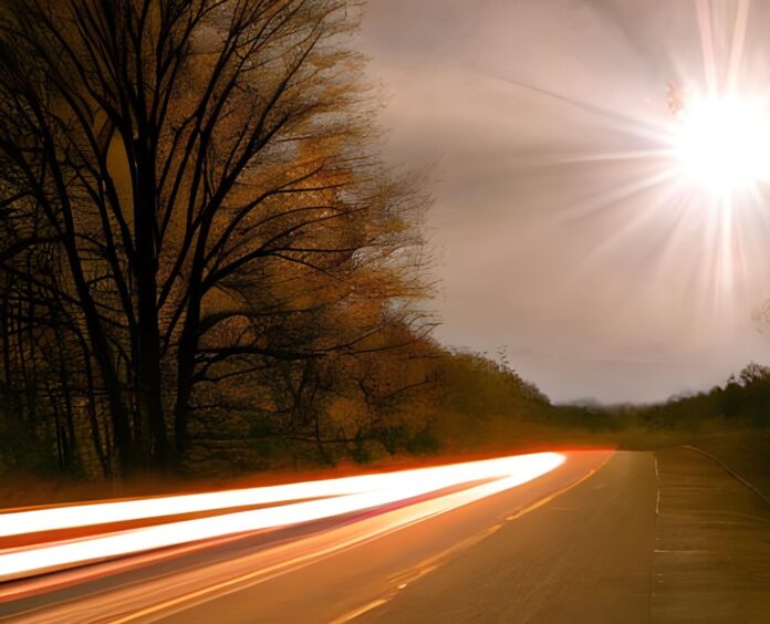 A low sun shines on a streak of a headlight beam along a road