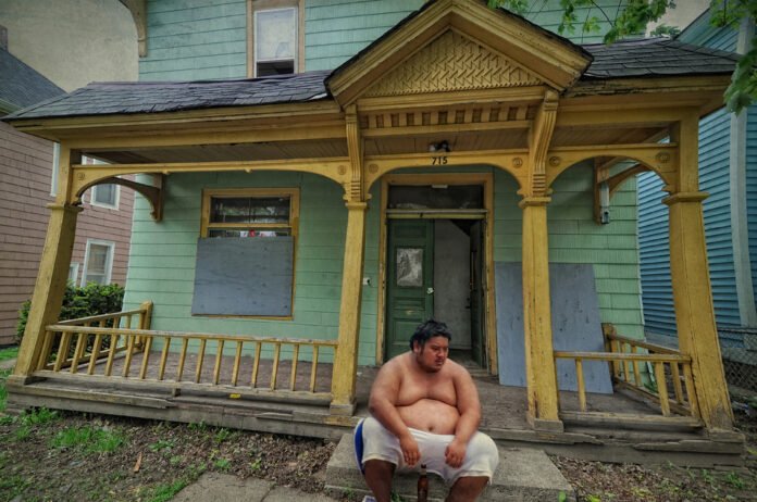 An older but brightly painted cottage with a heavyset man, naked from the waist up, sitting on the front steps
